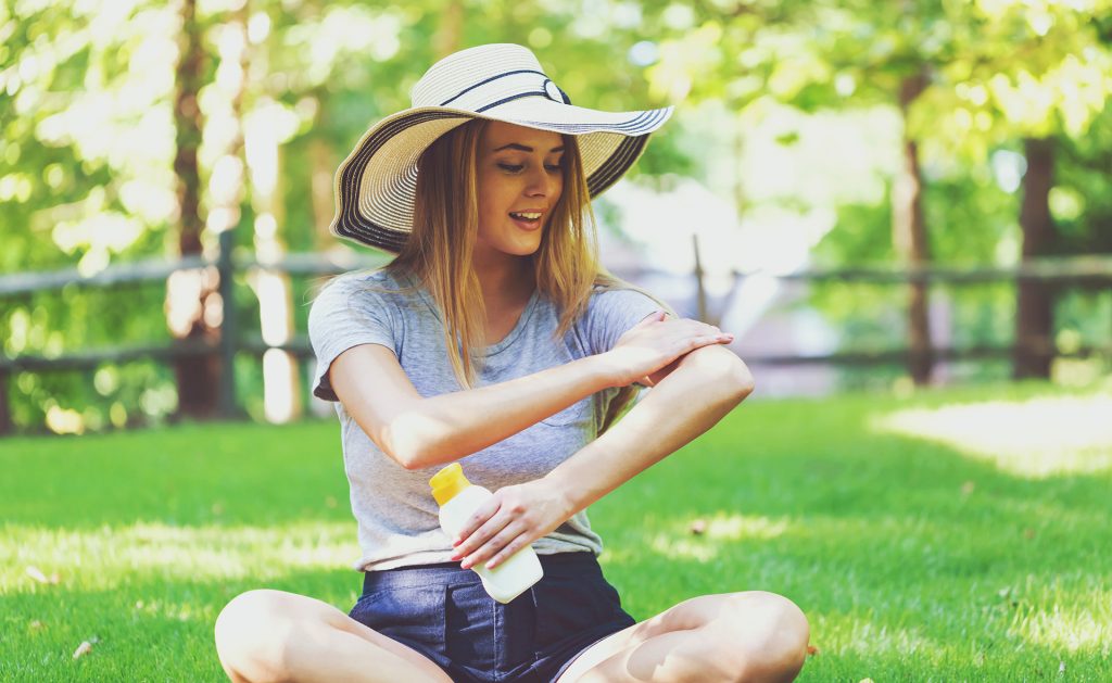 Woman in large sun hat on lawn applying sunscreen.