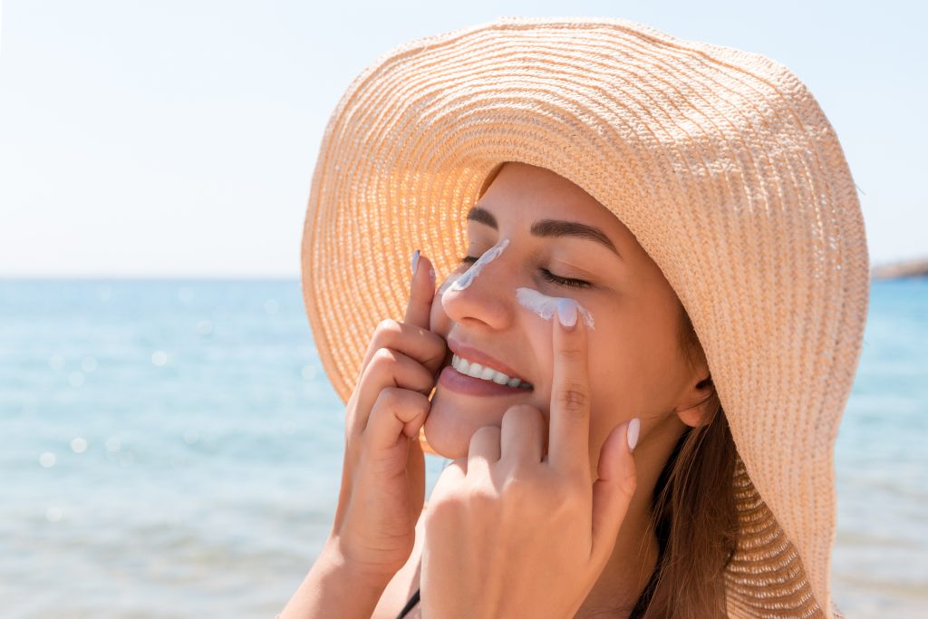 Woman at the beach in large sunhat applying sunscreen.