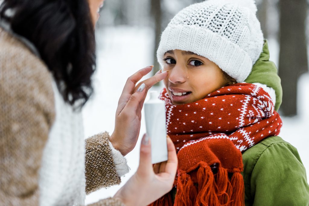 Mom applying sunscreen to daughter's nose in snowy setting.