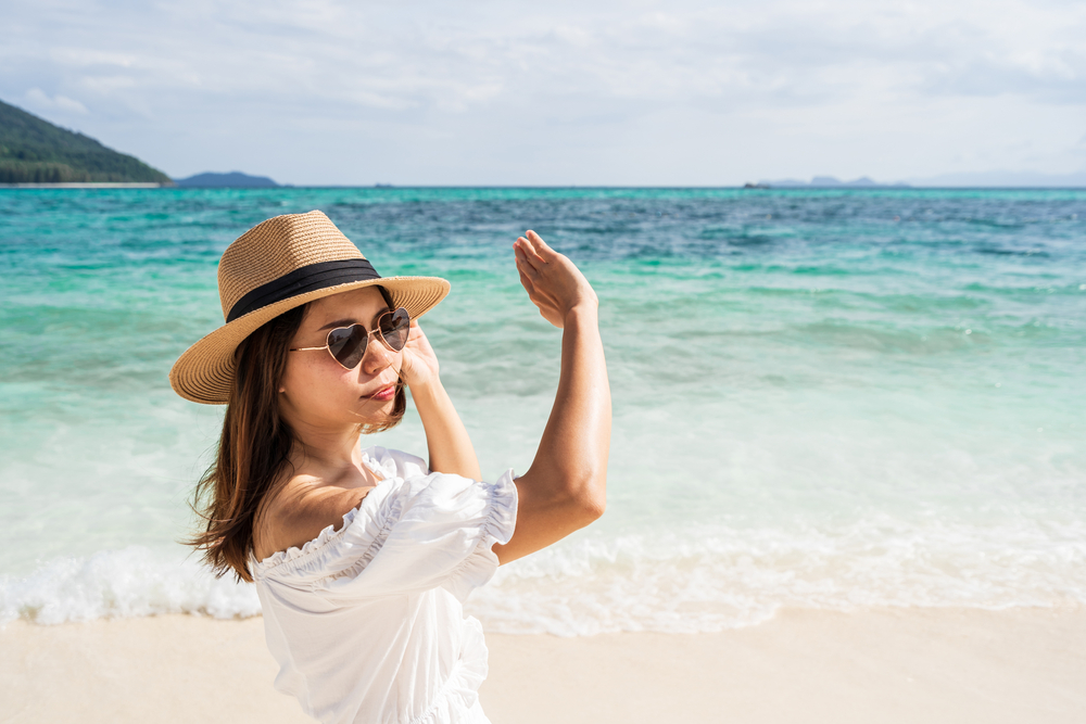 Woman wearing a sun hat and sunglasses protecting her skin from sun damage at the beach.