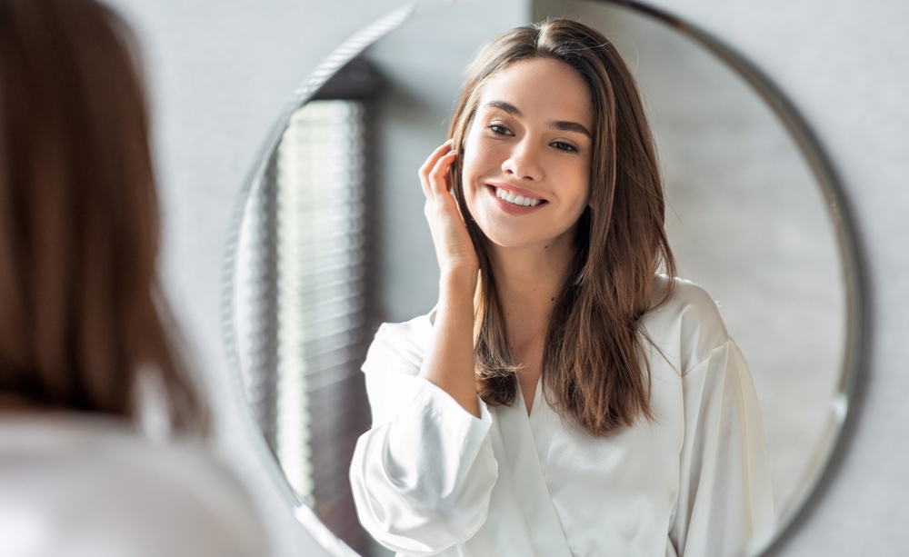 Woman smiling at her reflection with smooth, refreshed skin after collagen-stimulating Fraxel skin rejuvenation.